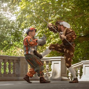 Deux escrimeurs en costumes renaissance s'affrontent sur une terrasse ombragée.