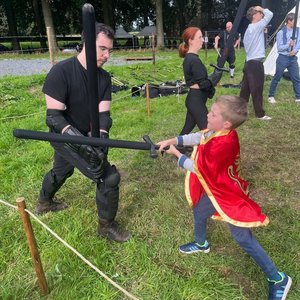 Enfant en cape rouge affrontant un adulte en tenue noire lors d’un combat d’épées en mousse.