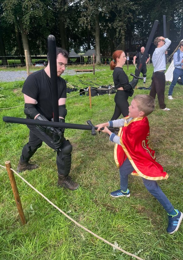 Enfant en cape rouge affrontant un adulte en tenue noire lors d’un combat d’épées en mousse.