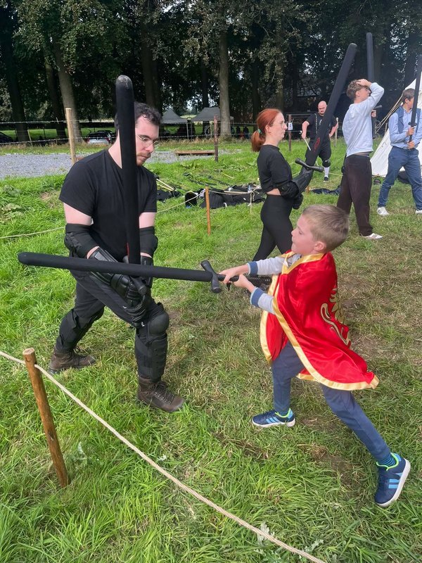 Enfant en cape rouge affrontant un adulte en tenue noire lors d’un combat d’épées en mousse.
