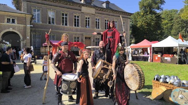 Groupe de musiciens et performers en costumes médiévaux, dont certains portent des tambours et des accessoires. Au fond, une belle architecture avec des fenêtres à guillotine et des tentes colorées pour un événement en plein air. Des visiteurs sont visibl