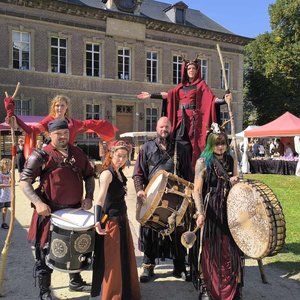 Groupe de musiciens et performers en costumes médiévaux, dont certains portent des tambours et des accessoires. Au fond, une belle architecture avec des fenêtres à guillotine et des tentes colorées pour un événement en plein air. Des visiteurs sont visibl