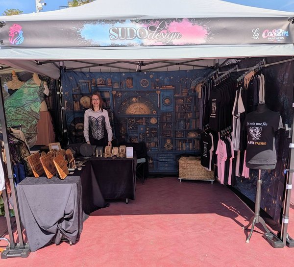 Stand sous tente SUDOdesign vendant T-shirts et objets artisanaux, exposante souriante.